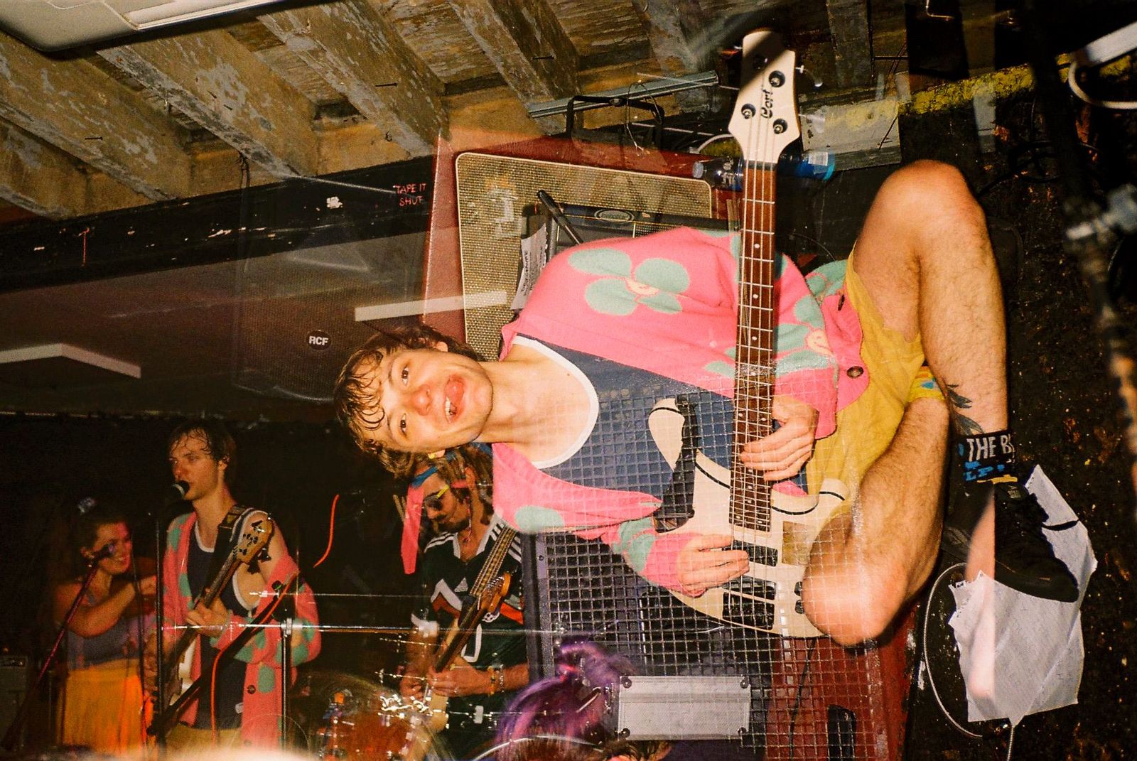 A bassist in a pink cardigan hanging upside down mid-performance while the band plays behind him.